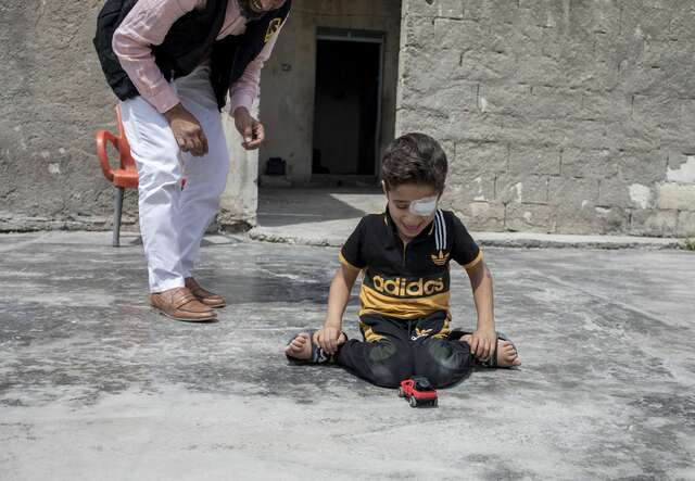 Ein Kind sitzt auf einem grauen Boden im Freien und spielt konzentriert mit einem Spielzeugauto, während ein Erwachsener in der Nähe steht und zusieht.